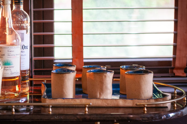 Six J Schatz ceramic tumblers with raw clay exteriors and dark glazed rims, on a brass bar tray beside spirit bottles